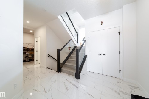 White marble-look tile flooring, glass and dark wood-finish staircase railing, recessed lighting, and white paneled doors with matte black hardware - 19 Sturtz Place, Leduc, AB 