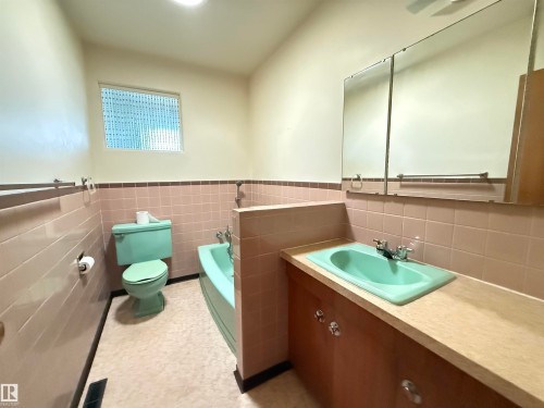 Spacious bathroom featuring dual vanities with wood-finish cabinetry and light-colored countertops, a soaking tub with turquoise tile surround, a separate toilet compartment, large mirror with integrated lighting, and natural light from a high window - 7007 91 Avenue Nw, Edmonton, AB 