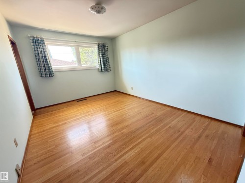 Bright bedroom featuring gleaming wood-finish flooring, white walls, and a generous window with plaid curtains allowing natural light throughout the space - 7007 91 Avenue Nw, Edmonton, AB 