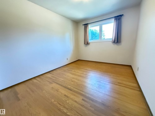Bright secondary bedroom featuring warm wood-finish flooring, clean white walls, and a large window with blue drapery providing natural light throughout the space - 7007 91 Avenue Nw, Edmonton, AB 