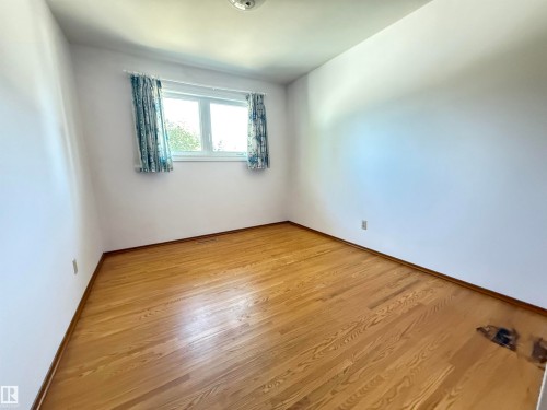 Bright secondary bedroom featuring gleaming wood-finish flooring, white walls, and a window with blue patterned curtains allowing natural light throughout the space - 7007 91 Avenue Nw, Edmonton, AB 