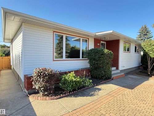 Modern single-story residence featuring horizontal siding in soft blue and burgundy accents, large picture windows with wood-finish frames, brick foundation detailing, and a welcoming entry with a burgundy front door - 7007 91 Avenue Nw, Edmonton, AB 