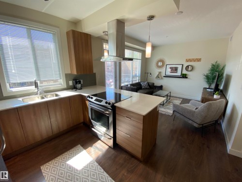 Open-concept living area featuring wood-finish flooring, a kitchen with stainless steel appliances, light wood-finish cabinetry, and solid surface countertops, illuminated by a pendant light fixture - #308 9907 91 Av Nw, Edmonton, AB 