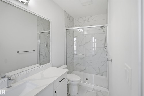 Vanity with integrated rectangular sink and chrome faucet, full-width wall mirror with overhead lighting, glass-enclosed shower with tiled surround, and white toilet - 2027 13 Avenue Nw, Edmonton, AB 