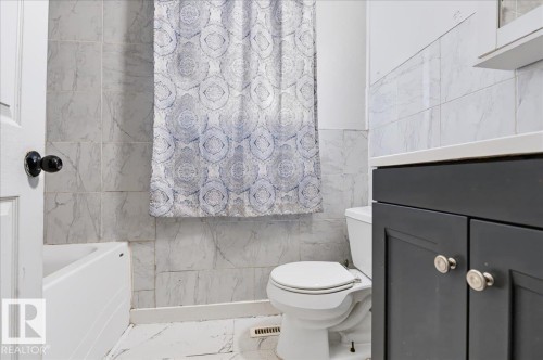 White tiled flooring, white bathtub, dark vanity with round pulls, white toilet, and tiled shower surround - 12919 135 Avenue Nw, Edmonton, AB 