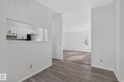 Open concept interior featuring wood-finish flooring, white walls, and a pass-through window to the kitchen with white cabinetry and a stainless steel refrigerator - 