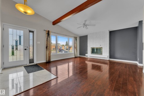 Vaulted ceiling with exposed wood beam, wood-finish flooring, white brick fireplace, bay window, and decorative glass entry door - 8803 68A Street Nw, Edmonton, AB 