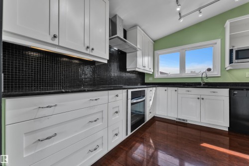 White cabinetry with dark countertops, black tile backsplash, stainless steel range hood, and wood-finish flooring - 8803 68A Street Nw, Edmonton, AB 