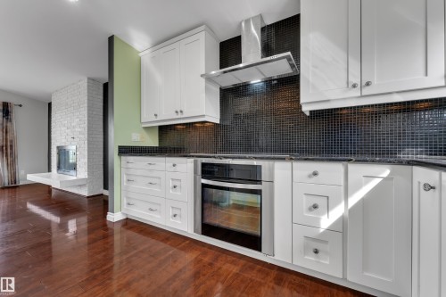 White shaker cabinetry with dark countertops, a black tile backsplash, stainless steel oven, and a built-in range hood - 8803 68A Street Nw, Edmonton, AB 