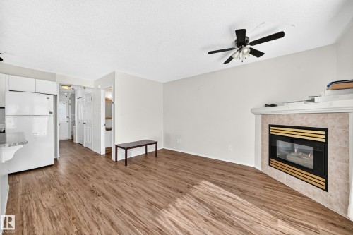 Wood-finish flooring throughout, featuring a gas fireplace with a tiled surround, a ceiling fan with light fixture, and white kitchen cabinetry with a refrigerator - #30 300 Hooper Cr Nw, Edmonton, AB 