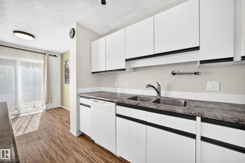 White cabinetry with dark hardware, dual basin stainless steel sink, integrated dishwasher, wood-finish flooring, and sliding glass door access - #30 300 Hooper Cr Nw, Edmonton, AB 