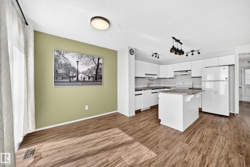 Bright open-concept kitchen and dining area featuring wood-finish flooring, white cabinetry, a central island with a stone-finish countertop, and a corner sink - #30 300 Hooper Cr Nw, Edmonton, AB 