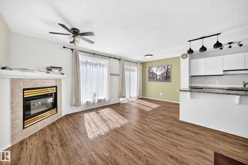 Open-concept living area featuring wood-finish flooring, a tiled fireplace with mantel, white cabinetry with a light-colored countertop, and a ceiling fan - #30 300 Hooper Cr Nw, Edmonton, AB 