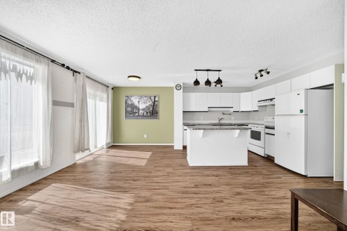 Open-concept living space featuring wood-finish flooring, a contemporary kitchen with white cabinetry, a kitchen island with seating, and a full suite of white appliances - #30 300 Hooper Cr Nw, Edmonton, AB 