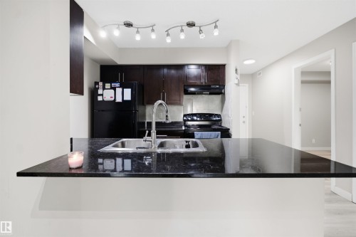 Kitchen featuring dark cabinetry, a black appliance package, a double basin stainless steel sink, and dark stone-finish countertops - #212 12035 22 Av Sw, Edmonton, AB 