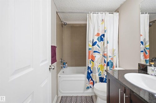 White panel door, tub-shower combination with tiled surround, dark wood-finish vanity with an undermount sink and dark countertop, and a wall-mounted mirror - #212 12035 22 Av Sw, Edmonton, AB 