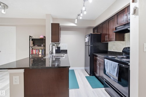 Kitchen featuring a dark stone countertop, dark wood-finish cabinetry, black appliances, and light wood-finish flooring - #212 12035 22 Av Sw, Edmonton, AB 