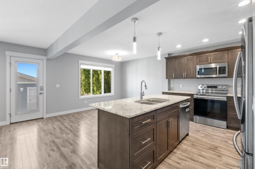 Open-concept kitchen featuring an island with a double basin sink, stainless steel appliances, wood-finish flooring, recessed lighting, and a glass-paneled exterior door - 2585 Pegasus Boulevard Nw, Edmonton, AB 