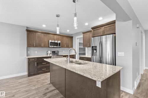 Kitchen featuring wood-finish cabinetry, stainless steel appliances, speckled stone countertops, and a center island with double sink - 2585 Pegasus Boulevard Nw, Edmonton, AB 