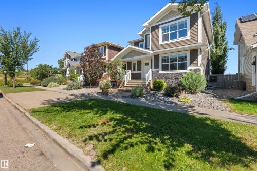 Two-story home with dark siding and stone-finish accents, featuring a front porch with white railings, a landscaped front yard with shrubs and gravel, and a concrete driveway - 2585 Pegasus Boulevard Nw, Edmonton, AB 