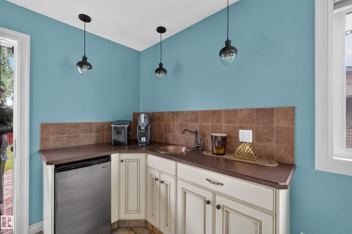 Corner kitchenette featuring a built-in beverage refrigerator, integrated sink with bronze-finish faucet, stone tile backsplash, and light-toned cabinetry - 131 Pine Drive, Wetaskiwin, AB - Indoor