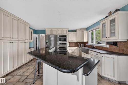 Spacious kitchen featuring a large island with dark speckled countertops, extensive cream-colored cabinetry, stainless steel appliances, a farmhouse sink, and tile flooring - 131 Pine Drive, Wetaskiwin, AB - Indoor Photo Showing Kitchen