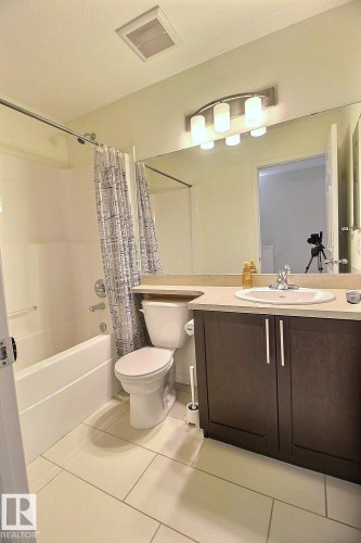 Built-in bathtub with shower, white porcelain toilet, dark wood-finish vanity, light-toned countertop with integrated sink, and contemporary light fixture - #18 4850 Terwillegar Cm Nw, Edmonton, AB 