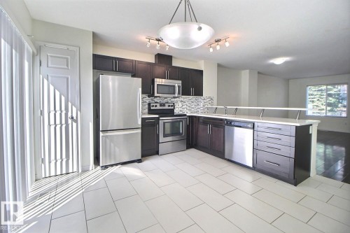 Kitchen featuring white tile flooring, dark wood-finish cabinetry, stainless steel appliances, a tile backsplash, and a breakfast bar with a white countertop - #18 4850 Terwillegar Cm Nw, Edmonton, AB 