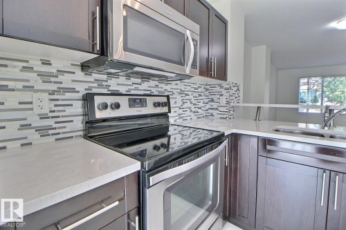 Kitchen featuring dark wood-finish cabinetry, light-toned countertops, and a full stainless steel appliance package, complete with a mosaic tile backsplash and undermount sink - #18 4850 Terwillegar Cm Nw, Edmonton, AB 