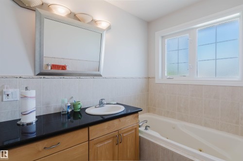 Built-in jetted tub, wood-finish vanity with an integrated sink, dark countertop, and a framed mirror with overhead lighting - 8517 156 Avenue Nw, Edmonton, AB 