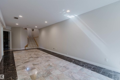 Recessed lighting illuminates the expansive room featuring a tiled floor with a dark perimeter border, light-toned walls, and a staircase with a wood handrail - 8517 156 Avenue Nw, Edmonton, AB 