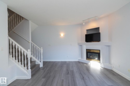 Corner fireplace with a black and brass-toned insert, wood-finish flooring, and a white painted staircase with traditional balusters - #37 3811 85 St Nw, Edmonton, AB 