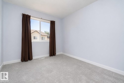 Bright secondary bedroom with neutral carpet flooring, white walls, and large window with brown thermal curtains - #37 3811 85 St Nw, Edmonton, AB 
