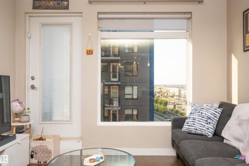 Living area featuring a large window with roller shades, a balcony door with frosted privacy glass, and dark wood-finish flooring - #610 5151 Windermere Bv Sw, Edmonton, AB - Indoor