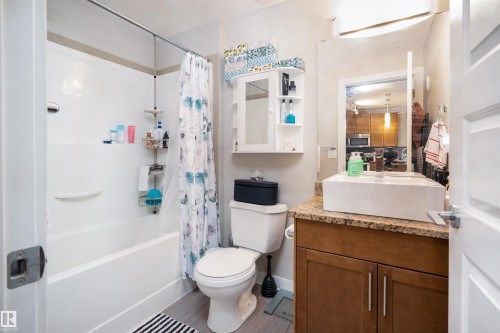 Bathroom featuring a white bathtub with shower, a vanity with a surface-mounted sink, a vanity mirror, wood-finish cabinetry, and a toilet - #610 5151 Windermere Bv Sw, Edmonton, AB - Indoor Photo Showing Bathroom