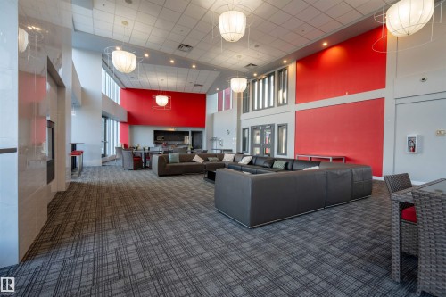 Spacious common area featuring high ceilings, contemporary pendant lighting, patterned carpeting, a full-height red accent wall, and extensive natural light from large windows - #610 5151 Windermere Bv Sw, Edmonton, AB - Indoor Photo Showing Other Room
