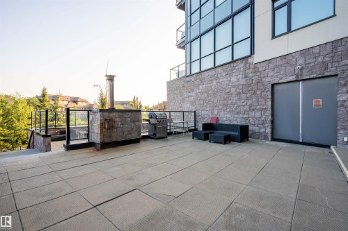 Spacious outdoor patio featuring stone-look paving, a stone-clad built-in structure with a metal chimney, and glass panel railings - #610 5151 Windermere Bv Sw, Edmonton, AB - Outdoor With Exterior