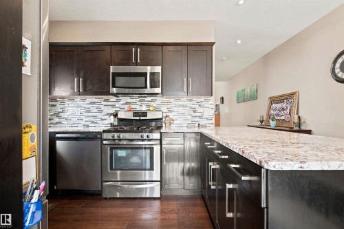Kitchen featuring stainless steel appliances, dark wood-finish cabinetry, a patterned tile backsplash, light-colored stone countertops, and recessed lighting - 10833 63 Avenue Nw, Edmonton, AB 