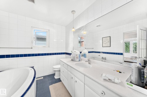 White tiled walls with a blue accent band, built-in bathtub, white vanity with integrated sink, and a large wall-mounted mirror - 3311 104 Avenue Nw, Edmonton, AB 