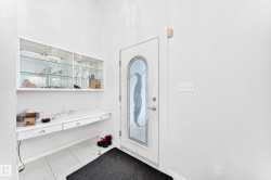 White entry door with decorative glass insert, white floating desk with drawers, white wall-mounted display cabinetry with glass doors, and light-toned tile flooring - 