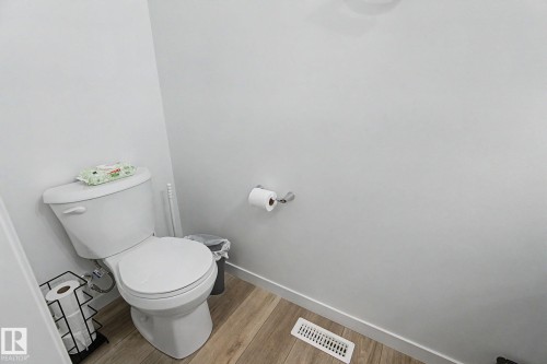 White toilet with chrome flush lever, wood-finish flooring, white baseboards, white walls, and a chrome toilet paper holder - #12 905 172 St Sw, Edmonton, AB - Indoor Photo Showing Bathroom