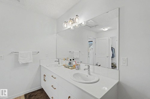 White dual vanity with integrated sinks and brass-finish hardware, illuminated by a contemporary light fixture, set against a full-width mirror - #12 905 172 St Sw, Edmonton, AB - Indoor Photo Showing Bathroom