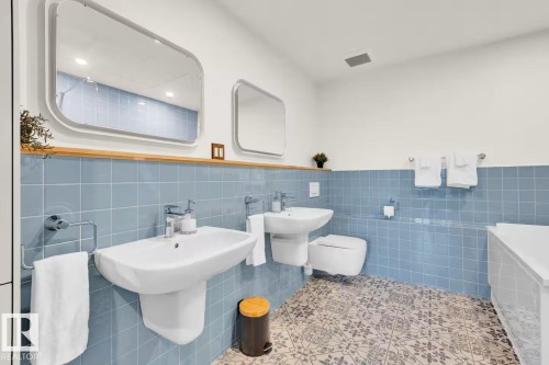 Twin rectangular mirrors, ceramic pedestal sinks, wall-mounted toilet, blue square tile wainscoting, and patterned floor tile - #903 10105 109 St Nw, Edmonton, AB 