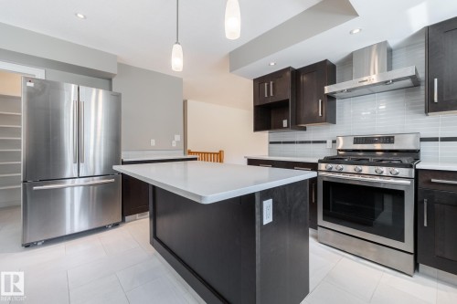 Modern kitchen featuring a central island with a light-toned countertop, dark wood-finish cabinetry, stainless steel appliances, and a tiled backsplash - 14 Highlands Place, Wetaskiwin, AB 