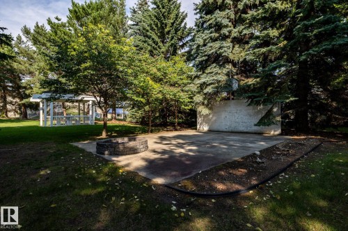 Concrete patio with a stone fire pit, surrounded by mature trees, a white gazebo, and a manicured lawn - 14 Highlands Place, Wetaskiwin, AB 