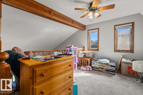 Sloped ceiling room featuring two windows with wood trim, a ceiling fan with integrated lighting, and a wood-finish railing - 8015 158 Ave Nw, Edmonton, AB 