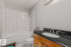 Bathtub with white tile surround, vanity with dark stone countertop, undermount sink, and wood-finish cabinetry - 