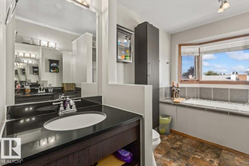 Vanity with dark countertop and undermount sink, large wall-mounted mirror, built-in soaking tub, and a window with wood-finish trim - 8015 158 Ave Nw, Edmonton, AB 