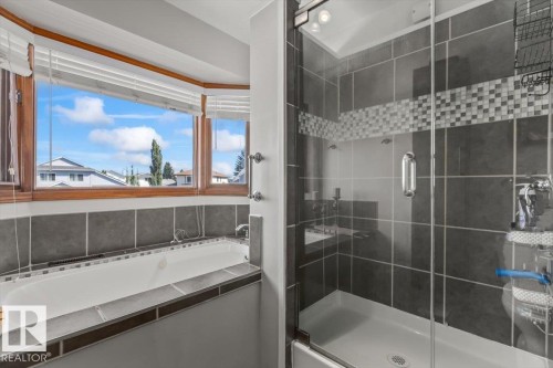 Built-in soaking tub with a tiled surround, wood-framed bay windows, and a glass-enclosed shower with dark gray tile and mosaic accents - 8015 158 Ave Nw, Edmonton, AB 