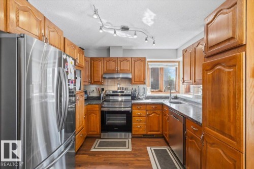 U-shaped kitchen featuring wood cabinetry, stainless steel appliances, dark countertops, and wood-finish flooring - 8015 158 Ave Nw, Edmonton, AB 